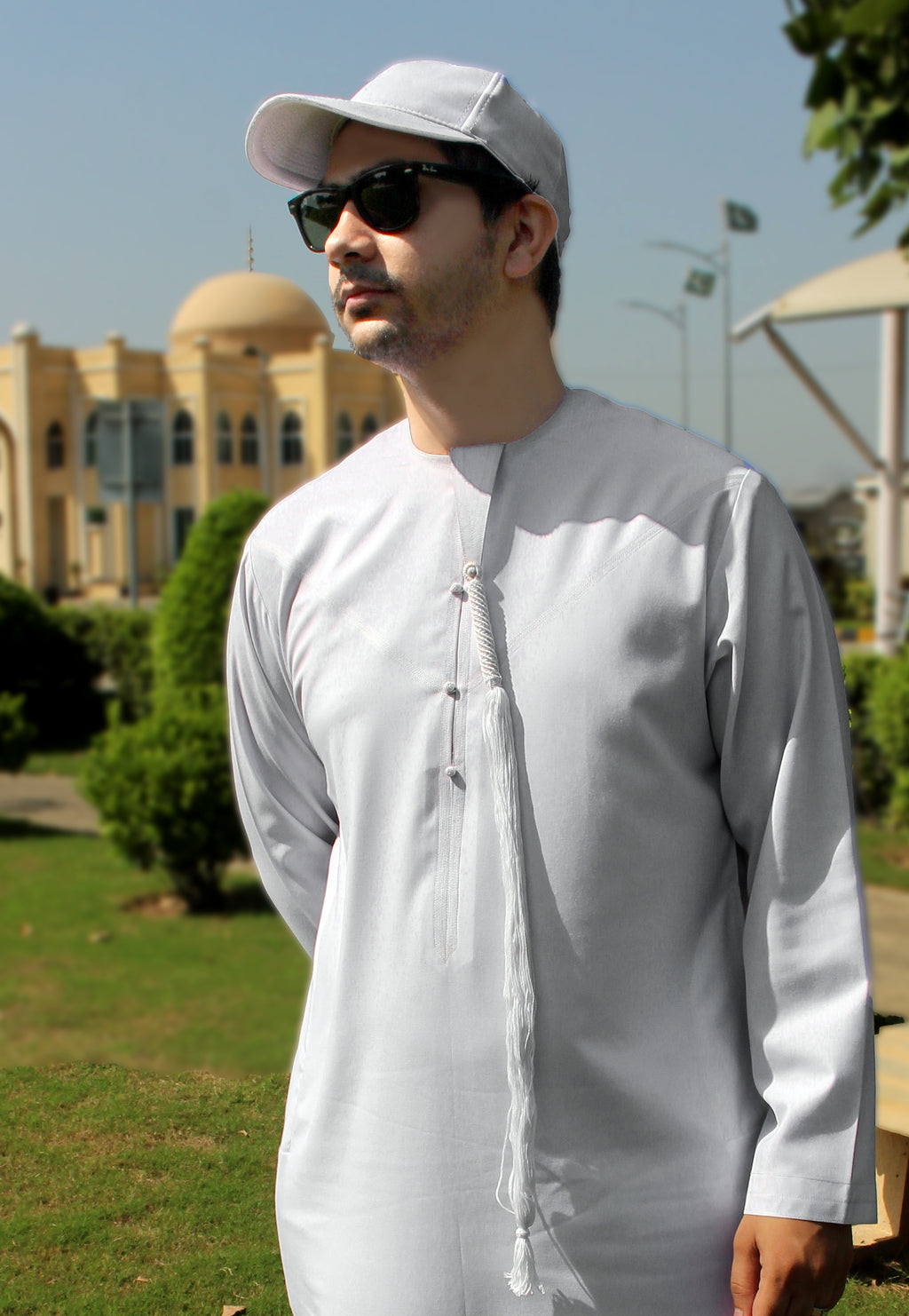 A man wearing a white long-sleeve traditional tunic (thobe) with a decorative front tie, a white baseball cap, and black sunglasses, standing outdoors in front of a domed building.