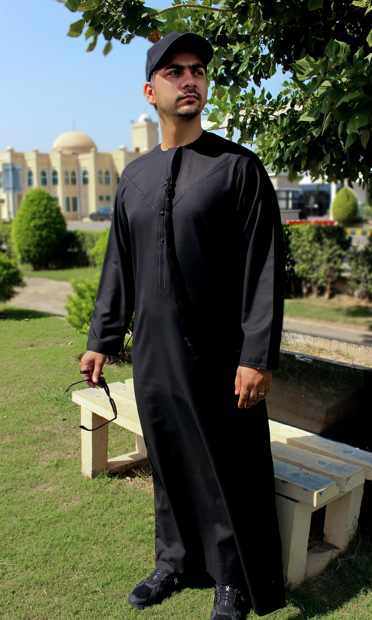 A man wearing a black thobe and black cap stands outdoors on a grassy lawn near a bench, with trees and a building in the background.