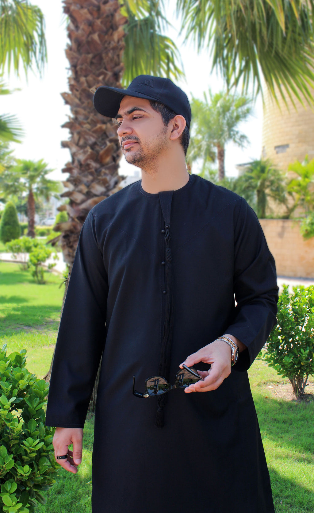 A man wearing a black long-sleeve thobe and black baseball cap, standing outdoors in a garden with palm trees and greenery in the background.