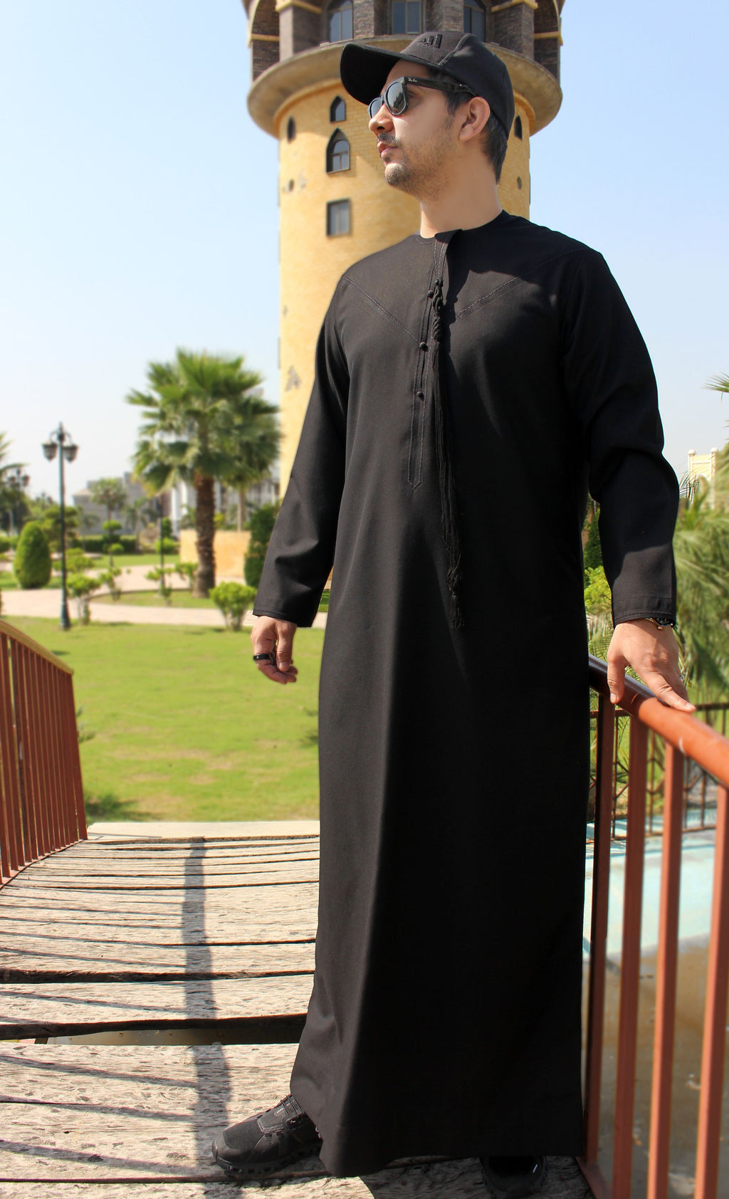 A man wearing a long black thobe and a black baseball cap stands on a wooden walkway outdoors with a tower, palm trees, and green lawn in the background.