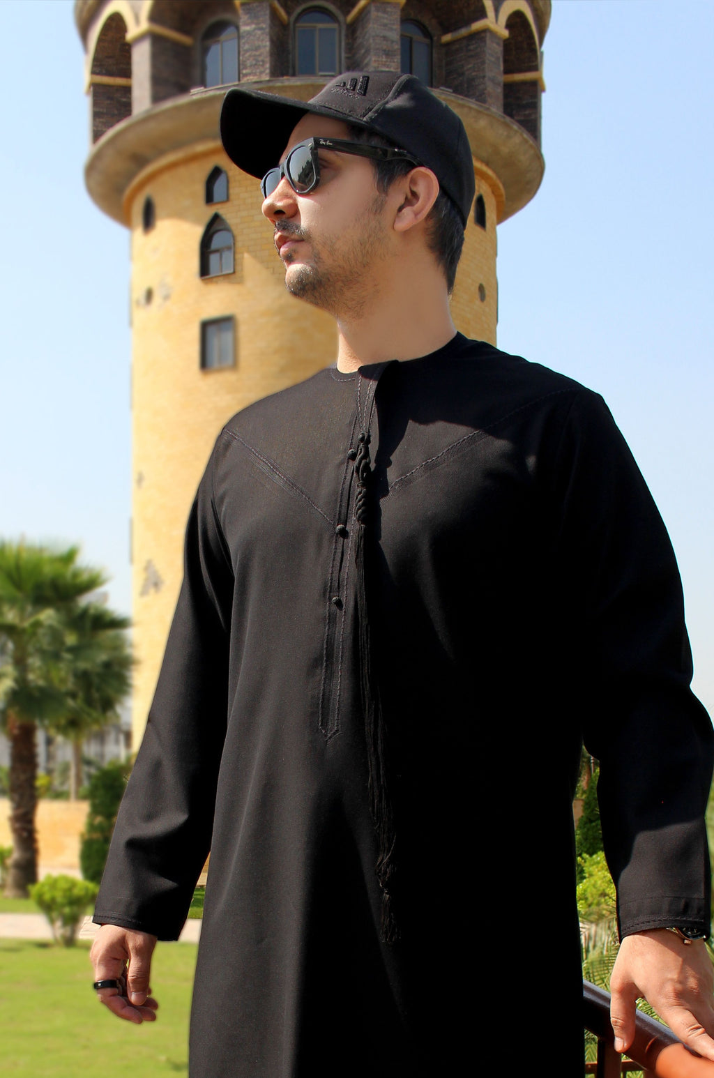 A man wearing a black long-sleeve thobe and a black baseball cap stands outdoors in front of a yellow tower under a clear blue sky.
