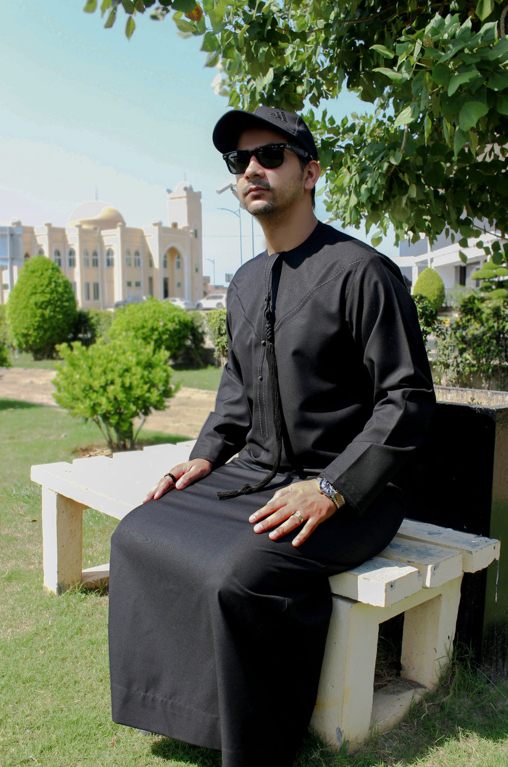 A man wearing a black traditional long-sleeve shirt and matching pants sits on a white bench outdoors under a tree, wearing a black cap and sunglasses.