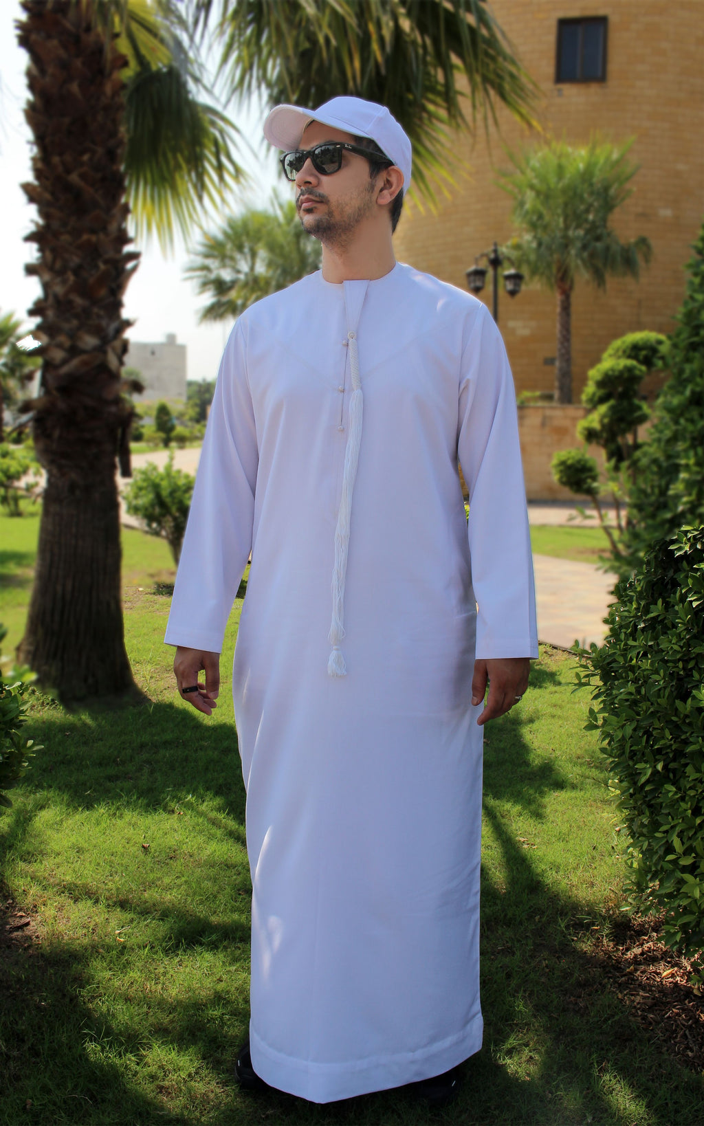 A man wearing a white traditional long robe and a white cap stands outdoors in a garden setting with palm trees and a building in the background.
