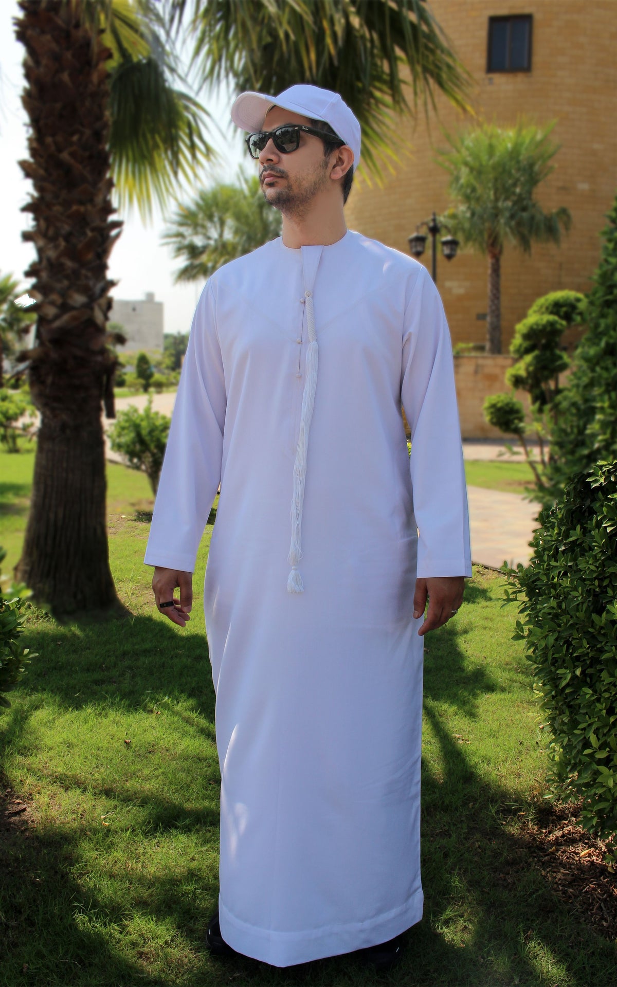 A man wearing a white traditional long robe and a white cap stands outdoors in a garden setting with palm trees and a building in the background.