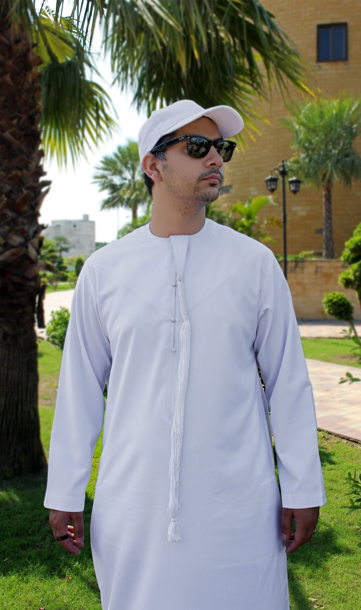 A man wearing a white long-sleeve thobe and white cap, standing outdoors in a garden setting with palm trees and a building in the background.
