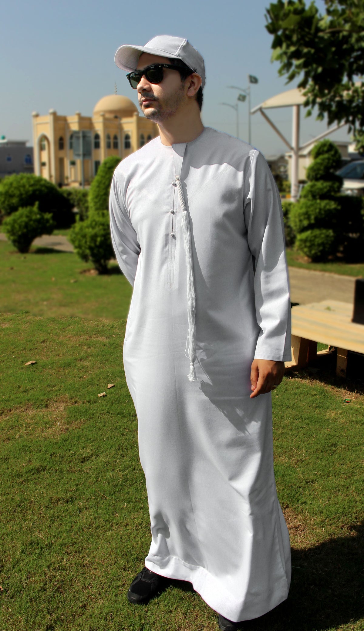 A man wearing a white long-sleeve thobe and matching white cap stands outdoors on a grassy lawn with trees and a domed building in the background.